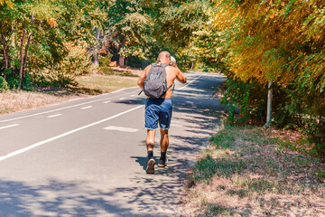 Tired aged sportsman in blue shorts and with a backpack behind his back walks after a jogging