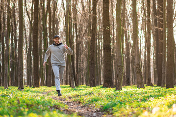 bearded young male wearing grey clothes does outdoors running outside in a sunny spring forest along pathway