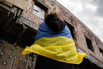 Woman with Ukrainian flag looking at abandoned destroyed building. Russia invasion, war in Ukraine, crisis concept. Girl standing in front of ruined, bombed residential house.