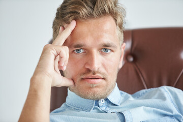 Fototapeta premium Portrait of a stylish intelligent man, small unshaven, charismatic, blue shirt, sitting on a brown leather chair, dialog, negotiation, short sleeve, brutal, hairstyle, emotions, energy, interest