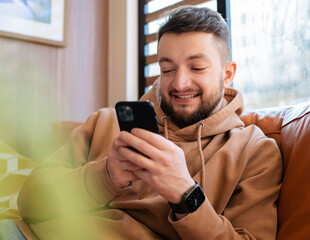 Smiling man holding phone, browsing apps, sitting on couch and spending leisure time with mobile device, chatting in social network.