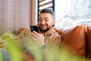 Fototapeta premium Smiling guy sitting on couch at cafe, using smartphone, chatting with friends or playing online. Happy man relaxing in living room, browsing internet or messaging with friends