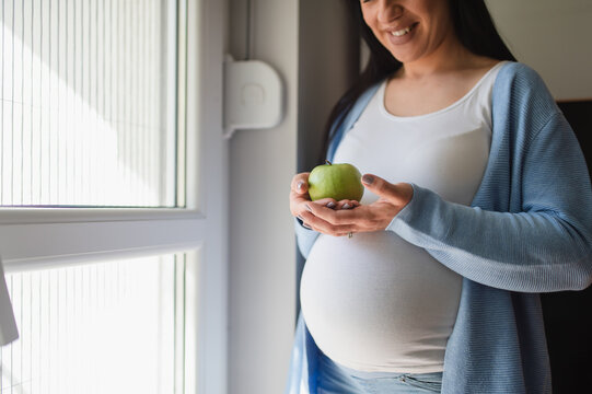 A smiling caucasian child-bearing woman stands next to the bright window and holds agreen apple with both her hands. Advanced pregnancy woman looks at apple. A black-hair female wears white t-shirt.  