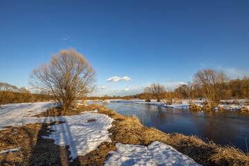 A picturesque landscape, early spring, a river with snow-covered banks, dry grass and bushes. March sunny day by the river. The first thaws, the snow is melting.