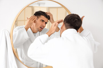 Worried young man with hair loss problem near mirror in bathroom