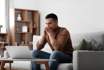 Troubled young man sitting on sofa at home - obrazy, fototapety, plakaty