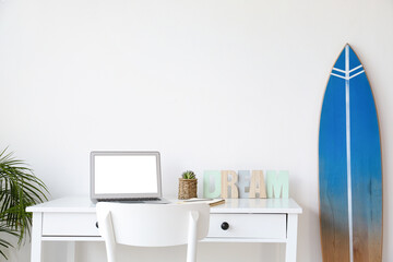 Workplace with modern laptop and surfboard near white wall