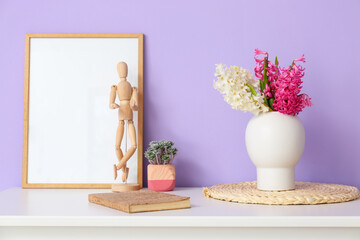 Blank photo frame, wooden mannequin doll, houseplant and vase with hyacinth flowers on table near color wall