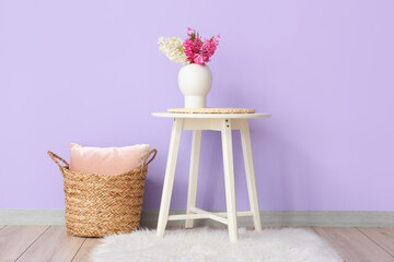 Vase with hyacinth flowers on table and wicker basket with pillow near color wall in room