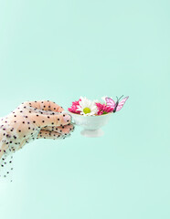 Spring creative arrangement of a woman's hand with a cup, flowers, and a butterfly on a blue background. The retro romantic aesthetic concept of the 80s and 90s. A surreal fashion idea for tea.