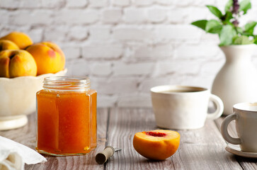 Homemade peach jam in a jar with some fresh peaches, two cups of coffee and a vase with green leaves, on a wooden table background. 