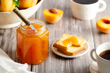 Homemade peach jam in a jar with some fresh peaches, two cups of coffee and some toasts, on a wooden table background. 