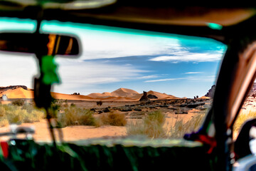 Windshield view from inside a car of Sahara desert sand dunes, rocky mountains mesa. Dry herbs and trees, colorful blue cloudy sky. Tadrart Rouge, Djanet, Illizi. Algeria.