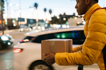 Black man with carton box riding scooter on street