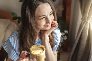 Happy cute woman with a cup of fragrant coffee in her hands is smiling and looking out the window...