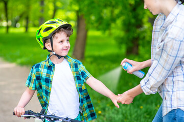 Mom spraying repellent on the skin of a smiling and looking son to protect against mosquitoes while cycling in the park. Close-up image