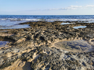 The coast of the Mediterranean Sea, long frozen lava, in the recesses of which there is sea water against a blue sky with clouds.