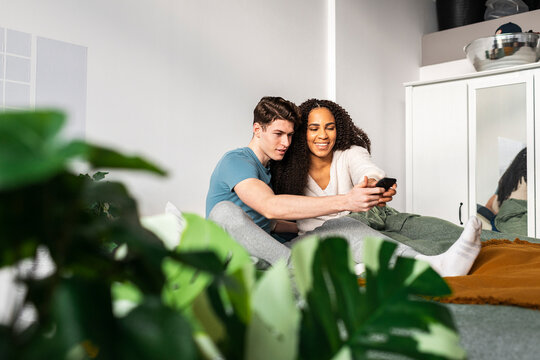 Multiracial couple browsing smartphone together on bed at home 
