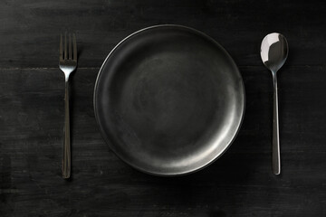 Modern black dining table setting with empty black plate, fork and spoon.