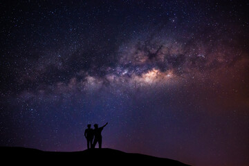 Naklejka na ściany i meble A couple standing on top of a mountain next to the Milky Way galaxy pointing at a bright star.