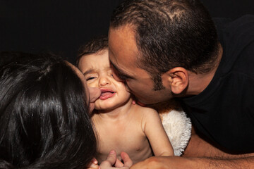 Young parents kiss their crying baby... Family portrait on black background. High quality photo