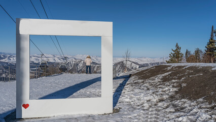 The frame for photographing is installed on a snow-covered high-altitude plateau. A man stands at the fence, looking into the distance, at the mountains. Cabs of the funicular on ropes. Altai.