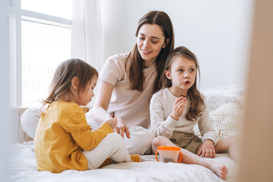 Young Happy Family With One Parent Woman Mother With Two Children Girls Eating Cookies On Bed Room At Home