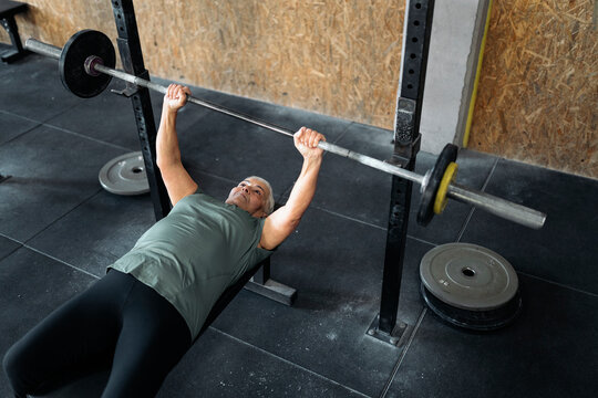 Elderly woman lying on bench and lifting barbell in gym
