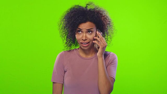 Young pretty curly African American woman gossip talking on phone discussing colleagues or tabloids gossiping and sharing news emotionally standing in green studio. Communication, people, emotions