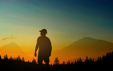 a silhouette of a person standing in front of a mountain range at sunset. The person is wearing a cap and appears to be looking out at the view.