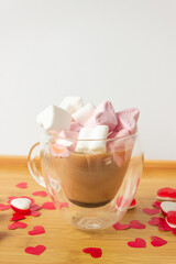 Glass double bottom cup of coffee with marshmallow in heart form. On the wooden table with red paper hearts and candies.
