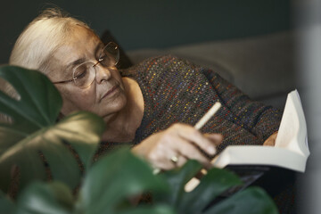 Mujer senior está leyendo un libro grueso en el sofá iluminada por una lámpara, con un lápiz...