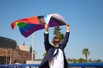 Mature gay man, executive, grey-haired, bearded, sunglasses, jacket and tie, waving the new lgbtiq+...