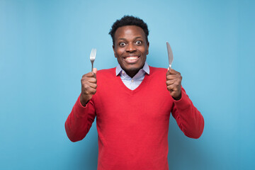 Hungry african young man holding fork and knife on hand ready to eat - obrazy, fototapety, plakaty