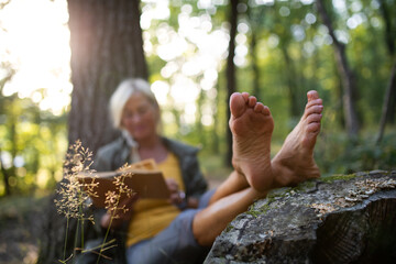 Senior woman relaxing and reading book outdoors in forest.