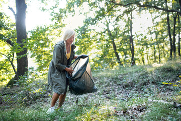 Senior woman ecologist with bin bag picking up waste outdoors in forest.