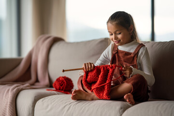 Little girl sitting on sofa and learning to knit indoors at home.