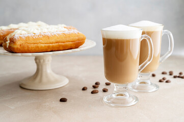 Donut or eclairs and coffee cups with whipped milk, on warm neutral background. Breakfast time.
