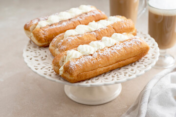 Donut or eclairs and coffee cups with whipped milk, on warm neutral background. Breakfast time.