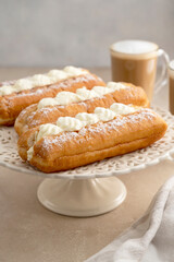 Donut or eclairs and coffee cups with whipped milk, on warm neutral background. Breakfast time.