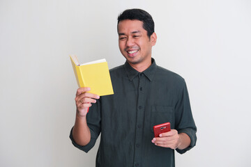 Adult Asian man smiling relax while reading a book and holding mobile phone - obrazy, fototapety, plakaty
