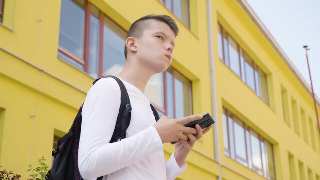 A Caucasian teenage boy works on a smartphone - view from below - a school in the background
