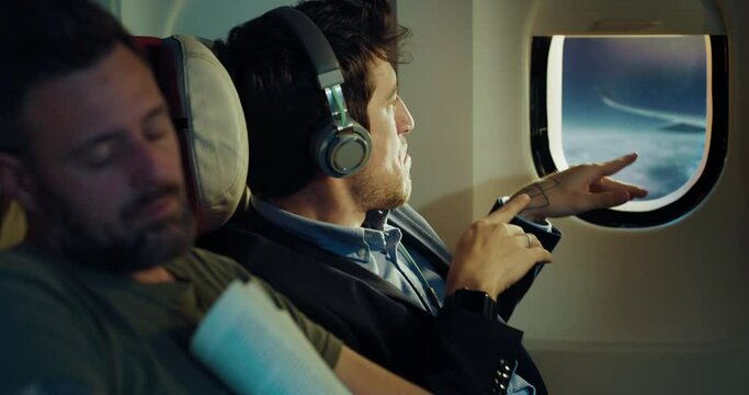 Cinematic shot of young businessman dances crazy while listening music and excuses for disturbing passenger sleeping near in aircraft while traveling with comfort in business class cabin during flight