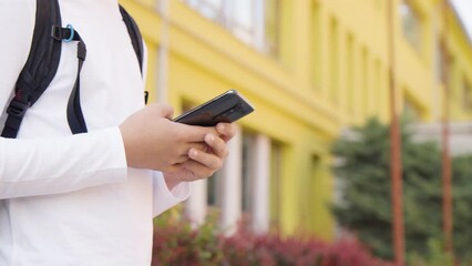 A Caucasian teenage boy looks at a smartphone - closeup on the phone - a school in the background - Powered by Adobe