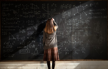 Cinematic shot of young female teacher is writing math formulas and equations with chalk on blackboard to explain educational material task to her pupils or students in college or university classroom - obrazy, fototapety, plakaty