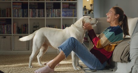 Cinematic authentic shot of young happy smiling woman is caressing and kissing with affection her lovely labrador retriever dog while having fun together on carpet in living room at home.