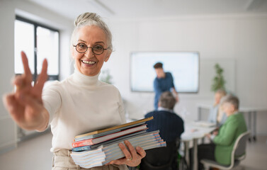 Happy senior woman student with books raising hand and looking at camera in classroom.