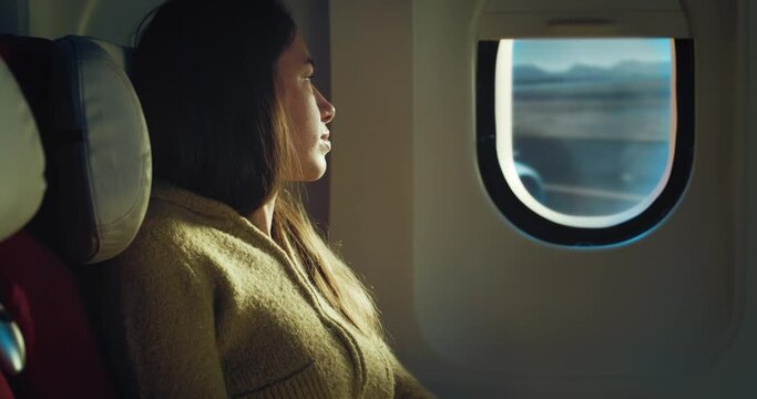 Cinematic shot of young woman sitting in cabin and looking out of window of aircraft in departure while traveling with comfort during international flight to vacation trip.