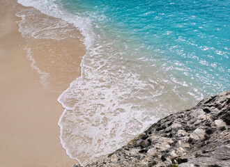 Sunny seascape with turquoise wave hitting white beige sandy coast and grey limestone rock. Colorful sea waters with white foam on sandy beach. Rock, sand and sea.