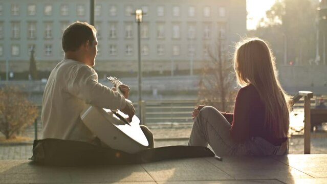 Romantic date on the waterfront. A couple of lovers are sitting on the embankment of the city. The guy plays the guitar for the girl.